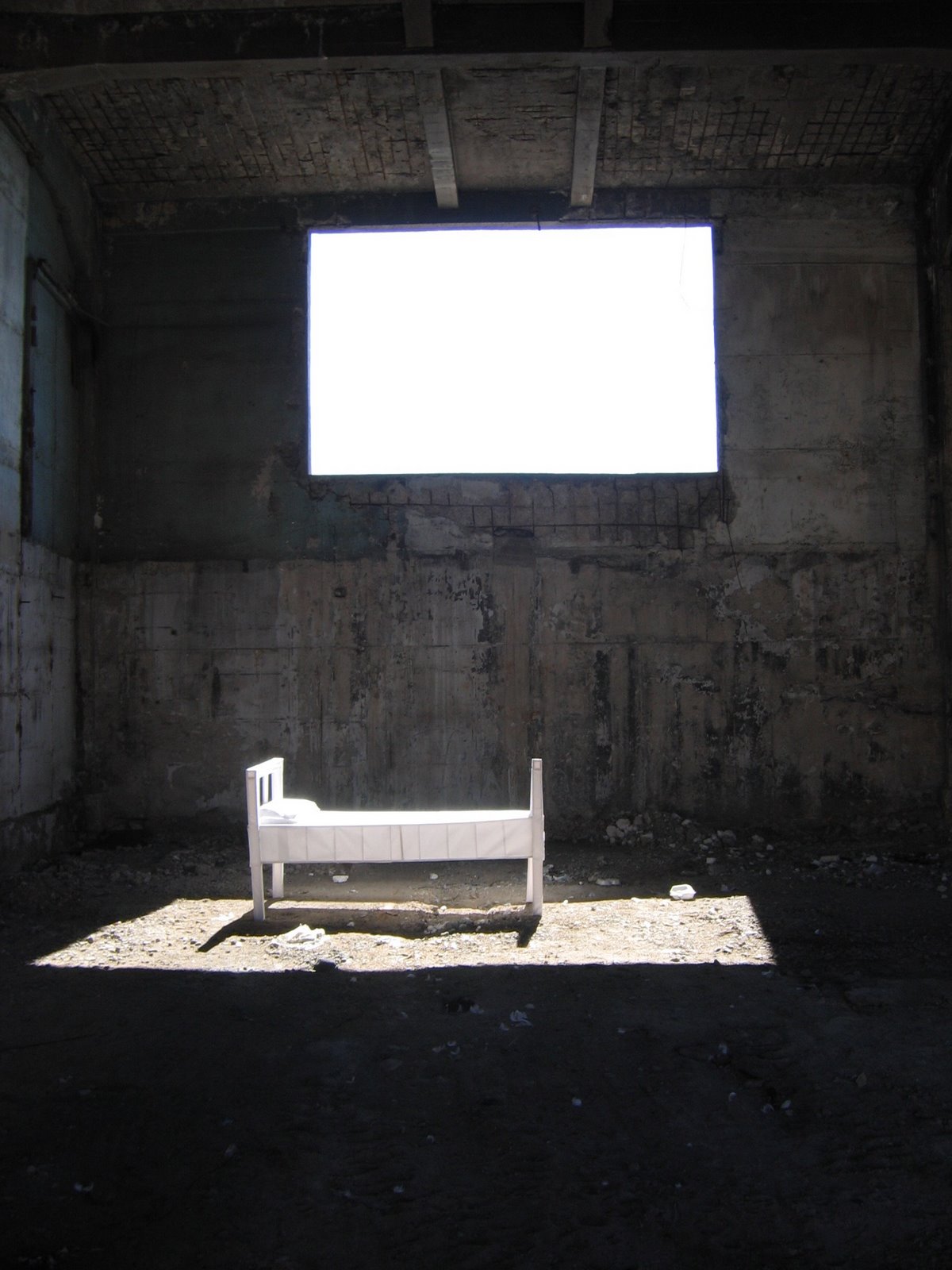 Cama blanca instalada en el interior de una vivienda en ruinas, bajo una ventana iluminada