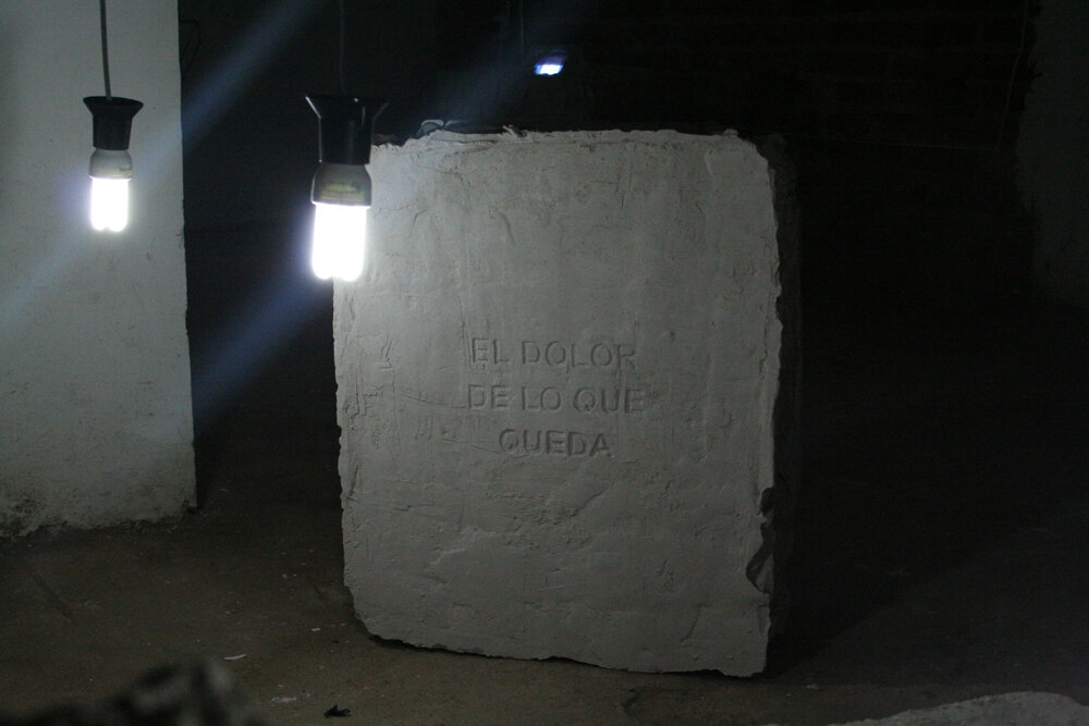 Bloque de yeso inscrito con el texto El dolor de lo que queda, iluminado por dos ampolletas colgantes